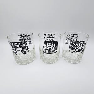 May include: Three clear drinking glasses with a textured base and black illustrations. Each glass features a different stylized figure. The glasses are set against a white background.