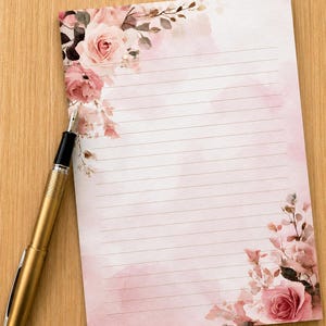 May include: A sheet of lined stationery paper featuring a soft pink watercolour wash and floral illustrations of roses in the top left and bottom right corners, accompanied by a metallic gold fountain pen with a black grip section.