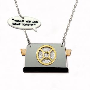 May include: A silver and black toaster necklace with a gold-coloured dial. The necklace has a white speech bubble charm that says "Would you like some toast?"
