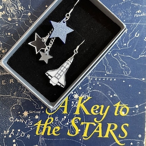 May include: A pair of silver earrings with a space shuttle charm and three star charms. The stars are black, silver, and blue. The earrings are in a small box with a blue background and the text "A Key to the Stars".