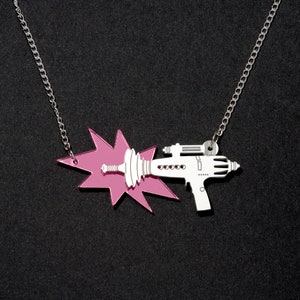 May include: A silver necklace with a laser gun pendant and a pink burst design. The laser gun is white with detailed accents. The necklace has a chain and a lobster clasp.