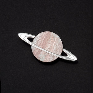 May include: A silver pin shaped like the planet Saturn with a pink and white ring system.
