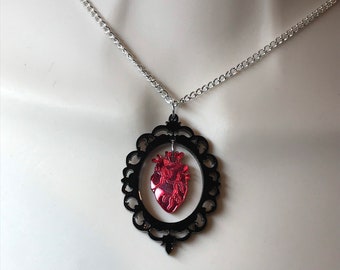 May include: A silver chain necklace with a black ornate frame pendant featuring a red anatomical heart.