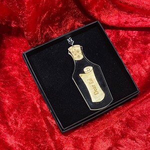May include: A clear glass bottle charm with a cork top and a rolled up scroll inside. The scroll has the words "Dear Ed" written on it.