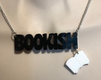 May include: A black acrylic necklace spelling "BOOKISH" with a small white book charm. The necklace hangs on a silver chain. The word "BOOKISH" is in a bold, sans-serif font.