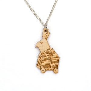 May include: A wooden necklace with a laser-cut pendant depicting a rabbit wearing a box with wheels. The rabbit is light brown and the box is a darker brown with a checkered pattern.
