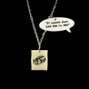 May include: A silver chain necklace with a white pendant featuring a black and white drawing of a creature. The pendant is attached to a white speech bubble that reads "It looks just like him to me!"