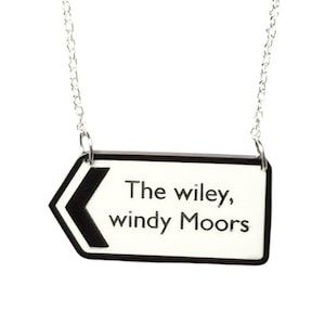 May include: A silver chain necklace with a pendant shaped like a road sign. The sign is white with black text that reads "The wiley, windy Moors" and a black arrow pointing left.