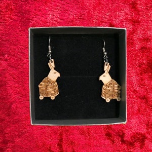 May include: A pair of wooden earrings shaped like a rabbit in a box on wheels. The earrings are silver and have a black background.