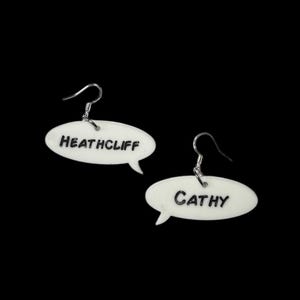 May include: White, speech bubble-shaped earrings with the names "HEATHCLIFF" and "CATHY" in black lettering. The earrings have silver-colored fish hook closures and are set against a black background.
