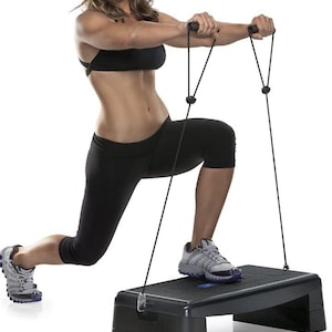 May include: A woman in black athletic wear using a black step platform with resistance bands. The platform is rectangular and has a textured surface. The woman is in a lunge position, holding the resistance band handles.