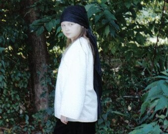 May include: A young person wearing a white shirt and black pants with a black bandana tied around their head. They are standing in a wooded area.