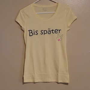 Puede incluir: Una camiseta amarilla con el texto "Bis später" y un conejito de dibujos animados con la lengua rosa en la parte delantera.