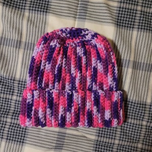 May include: A hand-knitted beanie hat in shades of pink, purple, and lavender. The hat has a ribbed design and a folded brim. The vertical stripes are created by the colour changes in the yarn. The hat is made of soft, textured yarn.