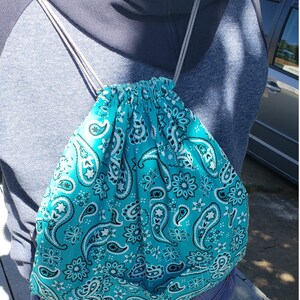 以下が含まれることがあります： ペイズリー柄のターコイズ色の巾着袋。青いパーカーと青いジーンズを着た人が持っています。
