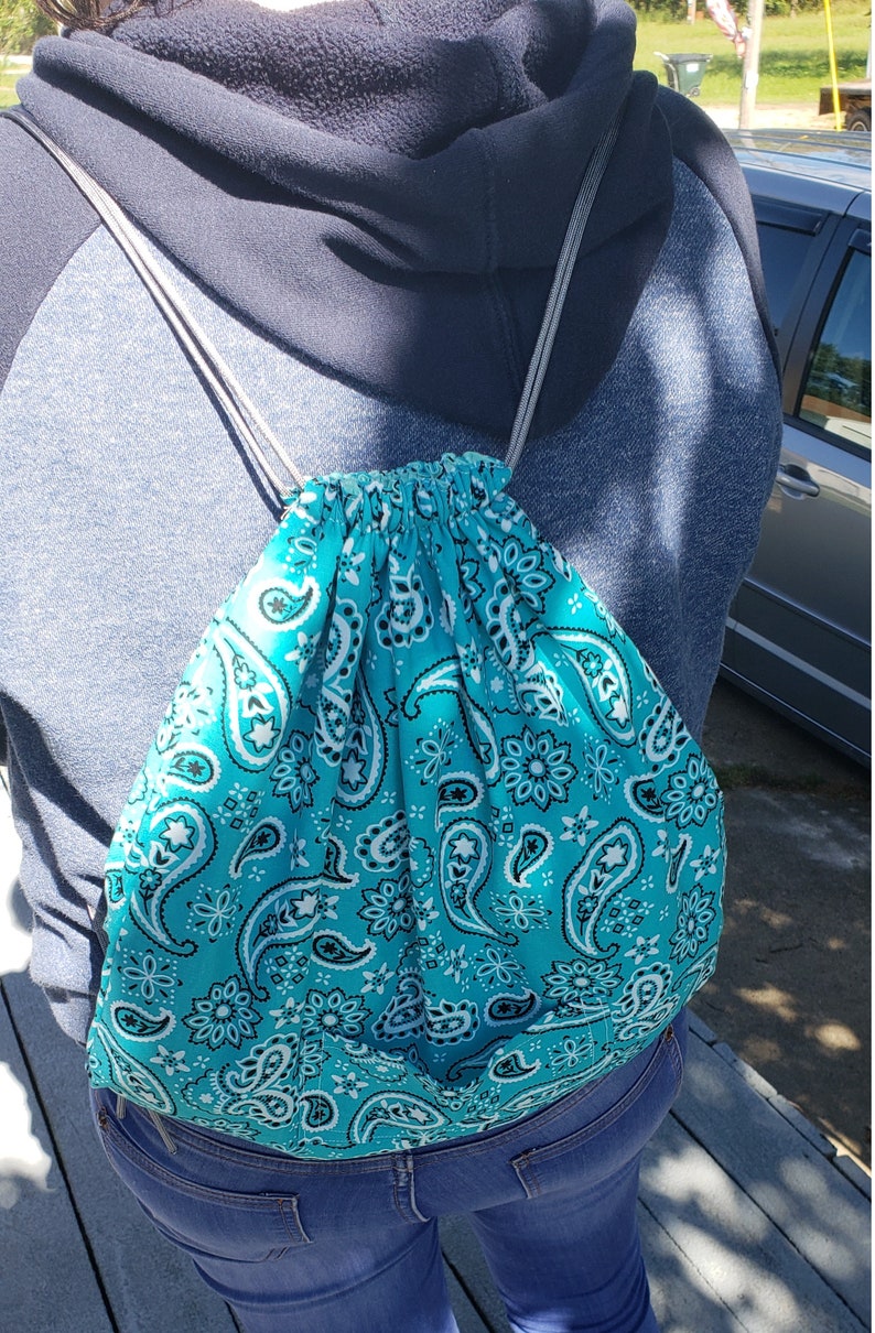 Puede incluir: Una bolsa de cord&oacute;n turquesa con un patr&oacute;n de paisley. La bolsa la lleva puesta una persona que lleva una sudadera con capucha azul y unos vaqueros azules.