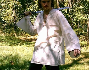 May include: A person wearing a white, long-sleeved shirt with a lace-up neckline, holding a sword in front of them. The person is standing in a grassy area with trees in the background.