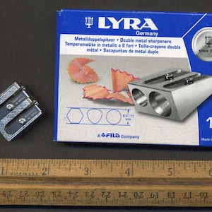 Puede incluir: Un sacapuntas doble de metal plateado con una caja azul. La caja tiene el nombre de la marca "LYRA" y el texto "Metalldoppelspitzer Double metal sharpeners" en blanco. La caja también tiene el texto "Made in China" y "since 1806".
