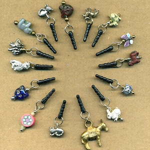 May include: A collection of 17 colourful and whimsical phone charms, each featuring a different design. The charms are attached to black phone plugs and include animals, insects, and abstract shapes.