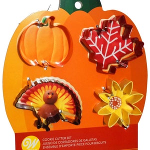 Puede incluir: Un juego de cuatro cortadores de galletas con forma de calabaza, hoja, pavo y girasol. Los cortadores son de metal y tienen un acabado brillante. El embalaje es naranja y presenta el logotipo de Wilton.