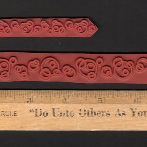 May include: Two red rubber stamps with a repeating pattern of circles and swirls. The stamps are approximately 2 inches long. The bottom stamp has the text "Do Unto Others As You Would Have" printed on a wooden ruler.