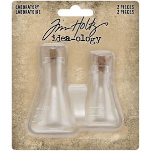 Pode incluir: Dois béqueres de laboratório de vidro transparente com rolhas de cortiça. Os béqueres são etiquetados com medidas em mililitros. Os béqueres são embalados em um recipiente de plástico transparente com o texto "Jim Holtz idea-ology" e "Laboratory Laboratoire" impresso na frente.