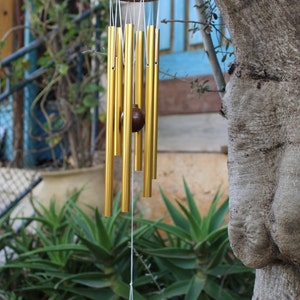 Peut inclure: Un carillon en or avec un sommet en bois et une étiquette en bois qui dit "Jerusalem Chimes". Le carillon est suspendu à une branche d'arbre.