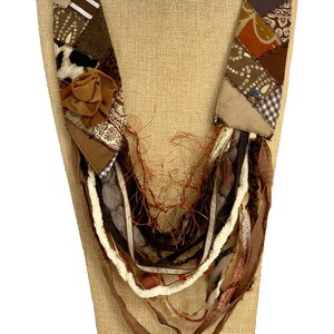 May include: A brown and beige patchwork fabric necklace with a brown fabric flower accent. The necklace is layered with brown, white, and beige fabric strips.