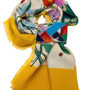 May include: A colorful floral scarf with a patchwork accent. The scarf features a vibrant mix of flowers in shades of red, purple, and blue on a white background. The edges are trimmed with yellow and a fringed border.