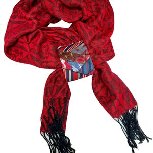 May include: A red scarf with a dark floral pattern and black fringe. A small, colorful patch is sewn onto the scarf. The scarf is made of a soft material and is suitable for cooler weather.