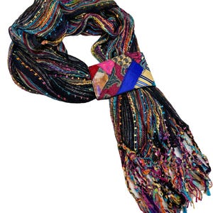 May include: A colorful scarf with a black base and vibrant threads of blue, orange, and pink. The scarf features a rectangular clasp with a patchwork design of various fabrics and colors. The ends of the scarf have a fringed trim.