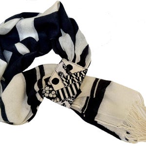 May include: A black and white scarf with a bold striped pattern. The scarf features a decorative clasp with geometric and floral designs. The end of the scarf has a fringe detail. The scarf is made of a lightweight material.