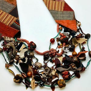 May include: A multi-strand necklace made of fabric scraps, beads, and wooden elements. The necklace features a variety of colors, including brown, red, green, and white. The fabric scraps are cut into different shapes and sizes, and the beads are made of different materials, including wood, glass, and metal.