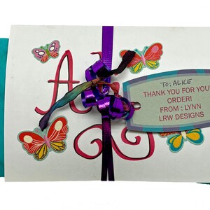 May include: A white card with the name "Alice" written in pink cursive. The card is decorated with colorful butterflies and a purple ribbon. A tag attached to the ribbon reads "To: Alice Thank you for your order! From: Lynn LRW Designs".