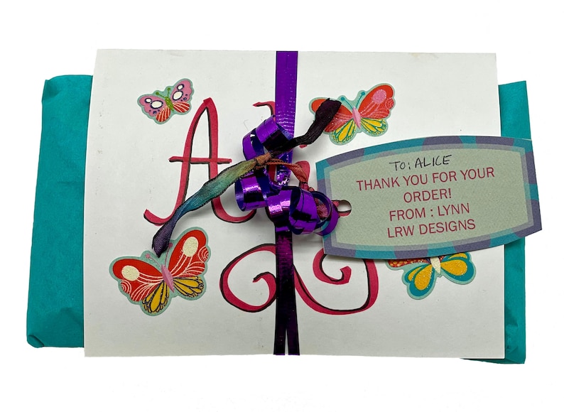 May include: A white card with the name "Alice" written in pink cursive. The card is decorated with colorful butterflies and a purple ribbon. A tag attached to the ribbon reads "To: Alice Thank you for your order! From: Lynn LRW Designs".