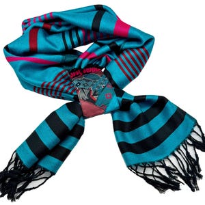 May include: A teal scarf with black and red stripes and fringed ends. The scarf has a patchwork design in the center with geometric patterns and a burgundy accent. The scarf is made of a soft, flowing material.