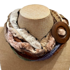 May include: A handcrafted infinity scarf in shades of brown, cream, and blue, displayed on a mannequin. The scarf features various textures, including fringe and a large wooden button closure. The design incorporates multiple fabrics and textures.