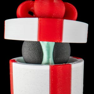 May include: A 3D-printed decorative gift box with a red bow on top. The box is white with red stripes and a gray sphere in the middle. A light green element is visible between the sphere and the box.