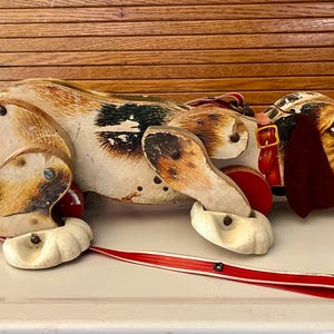 May include: Vintage wooden pull-along toy dog with a brown, white, and black spotted body. The dog has a red collar and a red and white pull string. The toy is on a white surface with a wooden background.