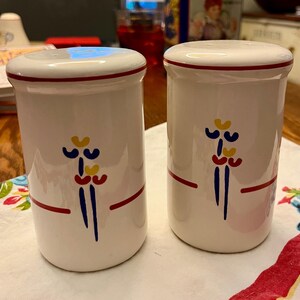 May include: A pair of cylindrical white ceramic salt and pepper shakers featuring a hand-painted abstract floral motif in red, blue, and yellow. Each shaker has a thin red horizontal band near the top and bottom, with a smooth, glossy finish.
