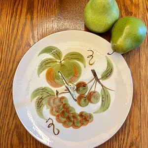 May include: A two-tiered serving tray with a white surface and a painted fruit design. The design includes green leaves, orange and green fruit, and a cluster of brown grapes. Two green pears are placed on the wooden surface next to the tray.