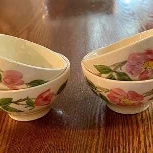 May include: Four cream-colored teacups with a floral design. Each cup has a pink flower and green leaves painted on the side. The handles are a dark green color. The cups are stacked in pairs.