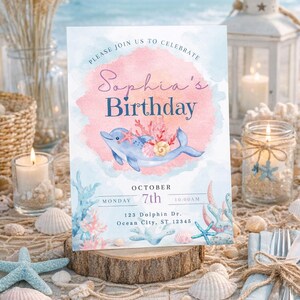 May include: A birthday invitation with a watercolor design featuring a blue dolphin, coral, and seashells. The invitation reads "Sophia's Birthday" and includes the date, time, and location. The invitation is set on a wooden slice with a beach theme.