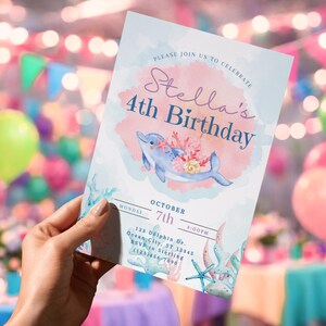 May include: A watercolor-style invitation for a 4th birthday party. The invitation features a blue dolphin with coral accents, the text "Stella's 4th Birthday," and event details. The background is a soft watercolor wash with a pink and blue color scheme.