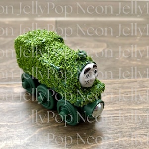 Puede incluir: Un tren de juguete verde cubierto de material verde texturizado, con una cara blanca y ruedas verde oscuro. El tren está sobre un fondo de madera. El texto "Neon JellyPop" es visible en el fondo.