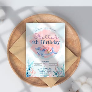 May include: A birthday invitation featuring a watercolor design with a dolphin, coral, and the text "Stella's 4th Birthday." The invitation is on a wooden tray with an envelope and cotton accents. The invitation includes the date and time.