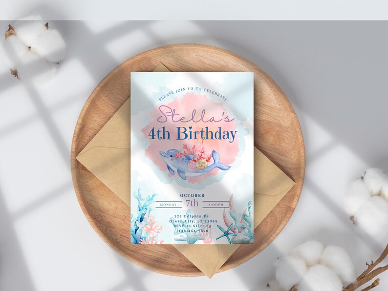 May include: A birthday invitation featuring a watercolor design with a dolphin, coral, and the text "Stella's 4th Birthday." The invitation is on a wooden tray with an envelope and cotton accents. The invitation includes the date and time.
