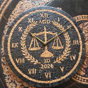 May include: A circular wall clock crafted from black and tan stone mosaic tiles. The clock face features Roman numerals, a central scale of justice icon, the letters AGD and JD, and the year 2026. Two thin gold-colored metal hands are centered.