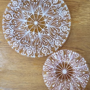 May include: Two clear glass plates with white floral designs. The plates have a circular shape with a central starburst pattern and ornate, swirling details. The plates are on a wooden surface.