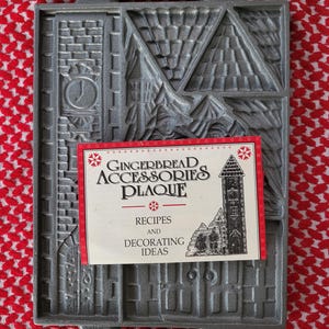May include: A rectangular gray cast metal gingerbread accessories plaque mold featuring embossed architectural details including a clock tower and roof sections. A white paper card with red borders and the text Gingerbread Accessories Plaque is placed on top.